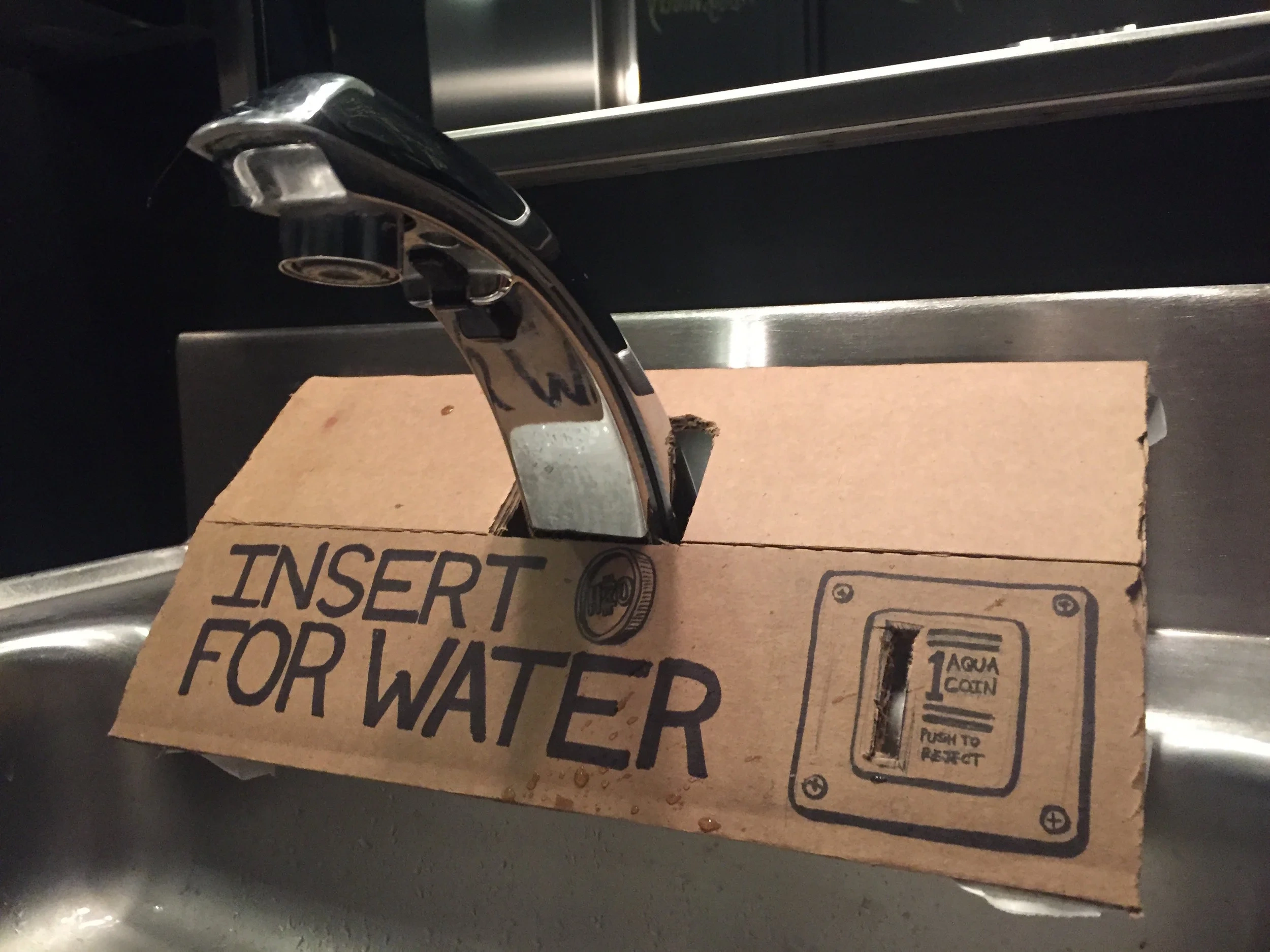 A cardboard coin-slot prototype reading “Insert for Water,” mocked up over a kitchen faucet